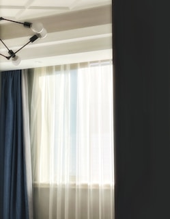 Sunlight filters through sheer white curtains, illuminating a room with elegant drapery on either side. A modern, geometric ceiling light fixture with exposed bulbs adds an artistic touch to the ceiling.