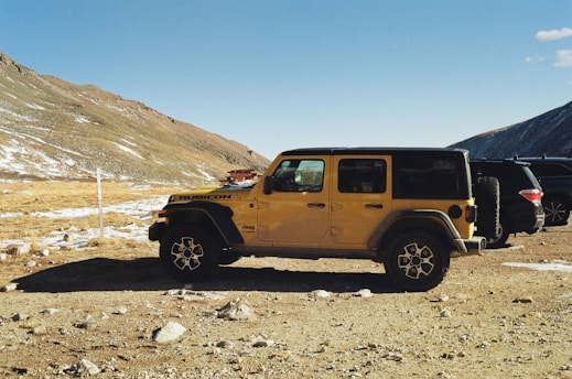 A yellow Jeep Rubicon is parked on a dirt area surrounded by a mountainous landscape. Some patches of snow are visible on the rugged terrain, and the sky is clear with a few clouds. Another vehicle is partly visible behind the Jeep.