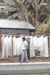 A person walks past a clothesline filled with white sheets that are drying outside. Another person in the background appears to be adjusting or hanging more sheets. The setting includes lush greenery with large leaves surrounding a small structure with a metal roof.