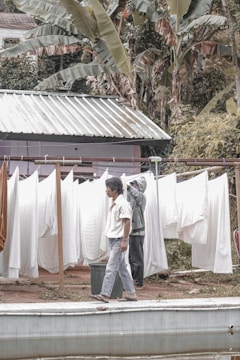 A person walks past a clothesline filled with white sheets that are drying outside. Another person in the background appears to be adjusting or hanging more sheets. The setting includes lush greenery with large leaves surrounding a small structure with a metal roof.