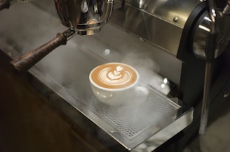 A steaming cup of coffee sits on a metallic tray under an espresso machine. The coffee has a beautifully crafted latte art design, with swirling patterns on top. The steam rises gently around the cup, indicating the freshness and warmth of the drink.