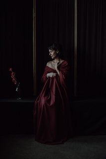 A person draped in a rich, deep red fabric sits against a dark curtain backdrop. A small vase with several red roses is positioned on a shelf to the left, adding a touch of color and contrast. The scene is dimly lit, focusing on the serene and contemplative expression of the individual.