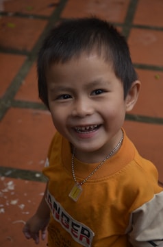 A young child with short dark hair, smiling brightly at the camera, wearing a yellow and orange shirt and a necklace with a small tag. The background consists of brown square tiles with green moss in the joints.