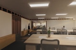 A modern, spacious office environment with wooden ceilings and large, collaborative tables. The room is lit by recessed lighting panels and features a minimalist decor with plants placed strategically around the space. There are a few people present, one standing near a cabinet and another seated at one of the desks, possibly working on a laptop.