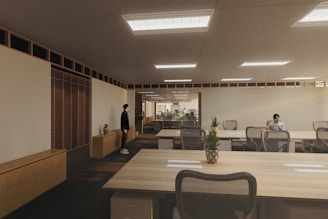 A modern, spacious office environment with wooden ceilings and large, collaborative tables. The room is lit by recessed lighting panels and features a minimalist decor with plants placed strategically around the space. There are a few people present, one standing near a cabinet and another seated at one of the desks, possibly working on a laptop.