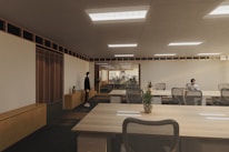 A modern, spacious office environment with wooden ceilings and large, collaborative tables. The room is lit by recessed lighting panels and features a minimalist decor with plants placed strategically around the space. There are a few people present, one standing near a cabinet and another seated at one of the desks, possibly working on a laptop.