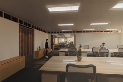 A modern, spacious office environment with wooden ceilings and large, collaborative tables. The room is lit by recessed lighting panels and features a minimalist decor with plants placed strategically around the space. There are a few people present, one standing near a cabinet and another seated at one of the desks, possibly working on a laptop.