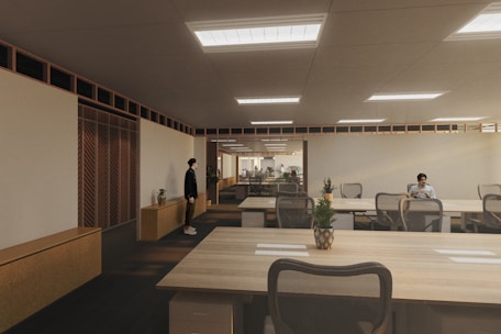 A modern, spacious office environment with wooden ceilings and large, collaborative tables. The room is lit by recessed lighting panels and features a minimalist decor with plants placed strategically around the space. There are a few people present, one standing near a cabinet and another seated at one of the desks, possibly working on a laptop.