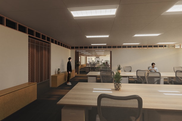 A modern, spacious office environment with wooden ceilings and large, collaborative tables. The room is lit by recessed lighting panels and features a minimalist decor with plants placed strategically around the space. There are a few people present, one standing near a cabinet and another seated at one of the desks, possibly working on a laptop.