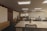 A modern, spacious office environment with wooden ceilings and large, collaborative tables. The room is lit by recessed lighting panels and features a minimalist decor with plants placed strategically around the space. There are a few people present, one standing near a cabinet and another seated at one of the desks, possibly working on a laptop.