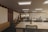 A modern, spacious office environment with wooden ceilings and large, collaborative tables. The room is lit by recessed lighting panels and features a minimalist decor with plants placed strategically around the space. There are a few people present, one standing near a cabinet and another seated at one of the desks, possibly working on a laptop.