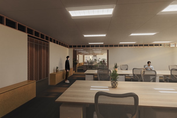A modern, spacious office environment with wooden ceilings and large, collaborative tables. The room is lit by recessed lighting panels and features a minimalist decor with plants placed strategically around the space. There are a few people present, one standing near a cabinet and another seated at one of the desks, possibly working on a laptop.