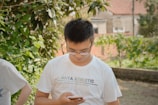 A person is standing outdoors in a garden-like setting, wearing a white t-shirt that has 'ANTA ATHLETIC' printed on it. The person is looking down at a smartphone they are holding. The background features green foliage and a blurred view of rustic buildings.