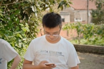 A person is standing outdoors in a garden-like setting, wearing a white t-shirt that has 'ANTA ATHLETIC' printed on it. The person is looking down at a smartphone they are holding. The background features green foliage and a blurred view of rustic buildings.