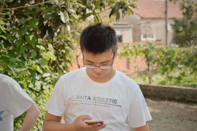 A person is standing outdoors in a garden-like setting, wearing a white t-shirt that has 'ANTA ATHLETIC' printed on it. The person is looking down at a smartphone they are holding. The background features green foliage and a blurred view of rustic buildings.