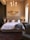 A stylish bedroom interior featuring a neatly made bed with white bedding, fluffy beige pillows, and a plush gray throw. The bed's headboard is tufted and flanked by two nightstands holding small elegant lamps. A decorative mirror with an ornate frame hangs above the bed. A modern chandelier with multiple light bulbs hangs from the ceiling, adding an elegant touch. An upholstered armchair with a studded finish sits in the corner. The room has a warm, neutral color palette with a mix of soft, cozy textures.