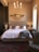 A stylish bedroom interior featuring a neatly made bed with white bedding, fluffy beige pillows, and a plush gray throw. The bed's headboard is tufted and flanked by two nightstands holding small elegant lamps. A decorative mirror with an ornate frame hangs above the bed. A modern chandelier with multiple light bulbs hangs from the ceiling, adding an elegant touch. An upholstered armchair with a studded finish sits in the corner. The room has a warm, neutral color palette with a mix of soft, cozy textures.
