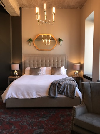 A stylish bedroom interior featuring a neatly made bed with white bedding, fluffy beige pillows, and a plush gray throw. The bed's headboard is tufted and flanked by two nightstands holding small elegant lamps. A decorative mirror with an ornate frame hangs above the bed. A modern chandelier with multiple light bulbs hangs from the ceiling, adding an elegant touch. An upholstered armchair with a studded finish sits in the corner. The room has a warm, neutral color palette with a mix of soft, cozy textures.