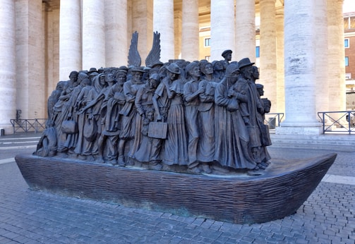 A bronze sculpture depicting a group of diverse people standing closely together on a boat. The figures represent different eras and cultures, each wearing period-specific clothing and carrying personal belongings. Behind them are large, white columns of a grand building.