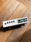 A small electronic device with multiple ports, including USB, ethernet, and audio jacks, resting on a wooden surface. The device is white with a rectangular shape and appears to be positioned at an angle.
