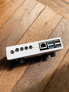 A small electronic device with multiple ports, including USB, ethernet, and audio jacks, resting on a wooden surface. The device is white with a rectangular shape and appears to be positioned at an angle.