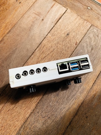 A small electronic device with multiple ports, including USB, ethernet, and audio jacks, resting on a wooden surface. The device is white with a rectangular shape and appears to be positioned at an angle.
