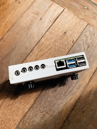 A small electronic device with multiple ports, including USB, ethernet, and audio jacks, resting on a wooden surface. The device is white with a rectangular shape and appears to be positioned at an angle.