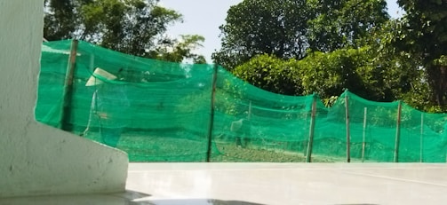 A green mesh fence stretches along a garden area with lush green trees in the background. The foreground features a smooth, shiny tiled surface, and part of a white structure is visible on the left side. Light and shadow patterns play across the scene, suggesting a sunny day.