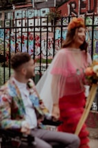 A couple stands in front of a colorful urban backdrop featuring graffiti on brick walls. The woman wears a bright, layered dress with sheer material and holds a bouquet, while the man is seated in a wheelchair wearing a vibrant jacket with a floral pattern. They appear happy and engaged in conversation.