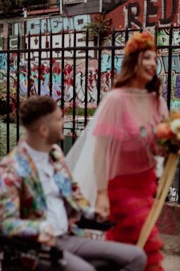 A couple stands in front of a colorful urban backdrop featuring graffiti on brick walls. The woman wears a bright, layered dress with sheer material and holds a bouquet, while the man is seated in a wheelchair wearing a vibrant jacket with a floral pattern. They appear happy and engaged in conversation.