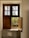 A wooden window with two sections is partially open, letting in light and revealing greenery outside. The upper part has frosted glass panes, while the lower section is a wooden shutter. The windowsill is tiled and reflects sunlight.