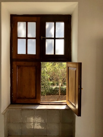 A wooden window with two sections is partially open, letting in light and revealing greenery outside. The upper part has frosted glass panes, while the lower section is a wooden shutter. The windowsill is tiled and reflects sunlight.