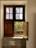 A wooden window with two sections is partially open, letting in light and revealing greenery outside. The upper part has frosted glass panes, while the lower section is a wooden shutter. The windowsill is tiled and reflects sunlight.