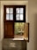 A wooden window with two sections is partially open, letting in light and revealing greenery outside. The upper part has frosted glass panes, while the lower section is a wooden shutter. The windowsill is tiled and reflects sunlight.