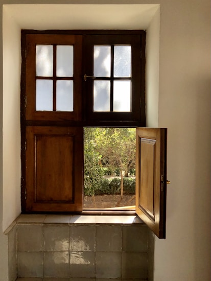 A wooden window with two sections is partially open, letting in light and revealing greenery outside. The upper part has frosted glass panes, while the lower section is a wooden shutter. The windowsill is tiled and reflects sunlight.