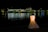 A glowing, dress-shaped lamp is positioned by a water body at night, with its reflection visible in the water. In the background, a row of illuminated buildings line the shore, casting colorful reflections across the rippling surface.
