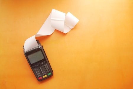 A black point-of-sale terminal with a paper receipt coming out of it, set against a solid orange background. The receipt paper is unrolled and cascades down to the side.