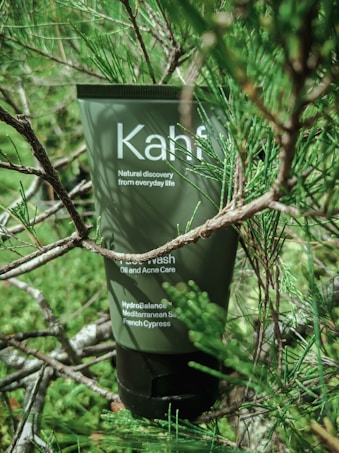 A green tube labeled 'Kahf' is nestled among lush green pine branches. The tube indicates it is a face wash for oil and acne care, with ingredients like Mediterranean sea and French cypress.