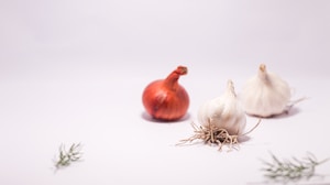 A red onion and two bulbs of garlic are positioned on a white surface with a light purple hue. Sprigs of rosemary are scattered around the vegetables, adding a touch of greenery to the scene. The composition is minimalist, with the focus on the natural textures and colors of the ingredients.