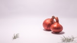 Two red onions with dried roots are placed on a clean, white surface. There are small sprigs of green herbs located in the bottom left and bottom right corners of the image.