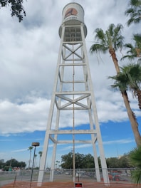 A tall, white water tower with a logo featuring a silhouette of a coyote on an orange background. The structure is constructed with a metal framework and is positioned prominently against a partly cloudy sky. Palm trees flank the sides, and there is fencing around the base of the tower. In the background, there are other trees and some buildings, possibly indicating an urban environment.