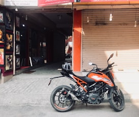 A parked motorcycle is positioned in front of a partially opened shutter, with sunlight casting shadows from nearby structures. The motorcycle has a sporty design with bright orange accents. A nearby shop displays framed artwork on its walls.