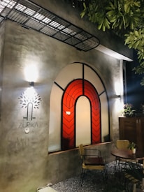 An outdoor patio area featuring a building wall with a large arched window adorned with geometric red stained glass panels. The wall is illuminated by wall-mounted lights, and a small round table with two chairs is positioned on gravel near the wall. Lush green leaves hang above, adding a touch of nature to the setting.