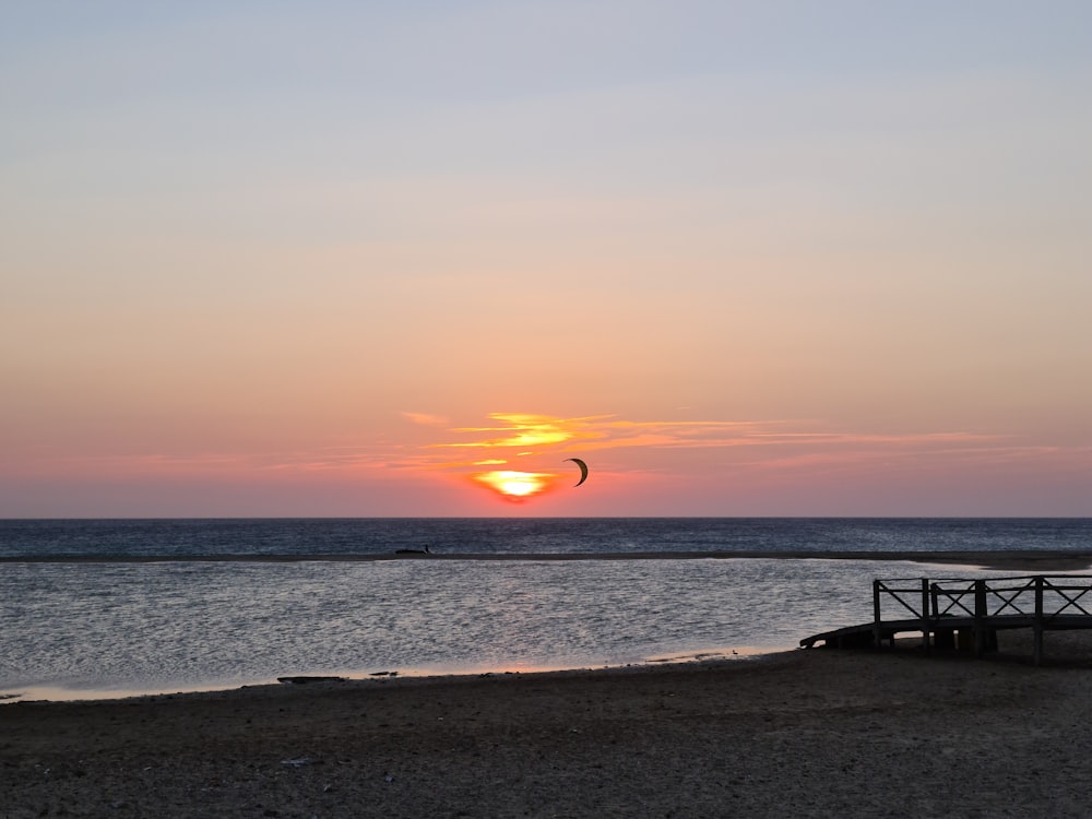 the sun is setting over the ocean with a kite flying in the sky