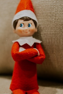 A small plush elf toy with a red and white outfit sits against a textured background. The elf has a mischievous expression with bright blue eyes, red clothing, and a white hat trim.