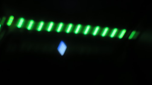 A blurred image featuring a series of glowing green lights arranged in a linear pattern across the top, with a singular glowing blue shape beneath them, set against a dark background.