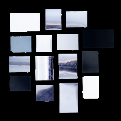 A collection of rectangular and square glass or acrylic panels arranged in a grid pattern against a black background. The panels exhibit a variety of textures and reflective surfaces, displaying gradients of grey, black, and white. Some panels appear to have a cloudy or marbled texture, adding to the visual interest.