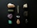 A collection of various gemstones and minerals arranged on a dark background. The stones vary in color and texture, with some displaying rough, uneven surfaces while others are smoother. Colors include hues of white, green, purple, black, and turquoise.