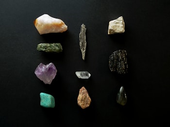 A collection of various gemstones and minerals arranged on a dark background. The stones vary in color and texture, with some displaying rough, uneven surfaces while others are smoother. Colors include hues of white, green, purple, black, and turquoise.