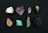 A collection of eight distinct mineral stones arranged in two rows against a dark background. The stones exhibit a variety of colors and textures, ranging from white to deep green and black, with some displaying a crystalline structure.